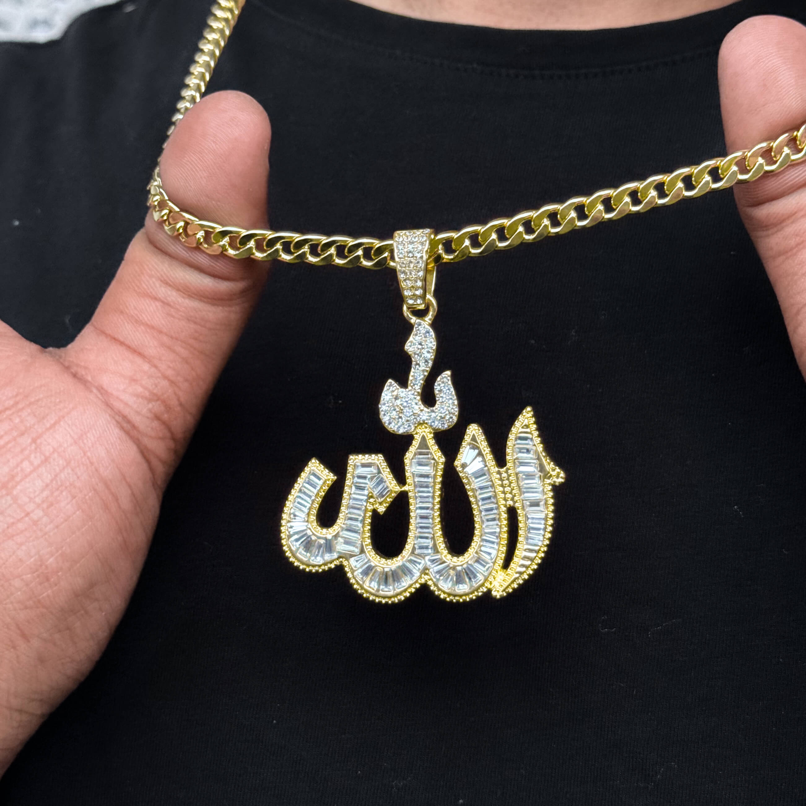 Close-up of gold baguette Allah pendant with 6mm Cuban chain held by hand for detail view.