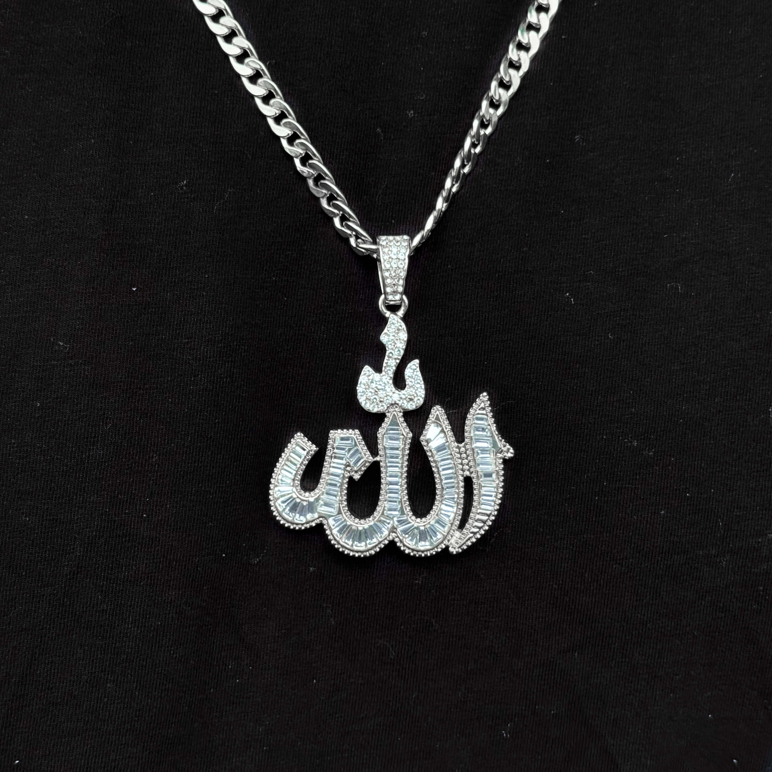 Detailed silver baguette Allah pendant with Cuban chain shown against black shirt background.