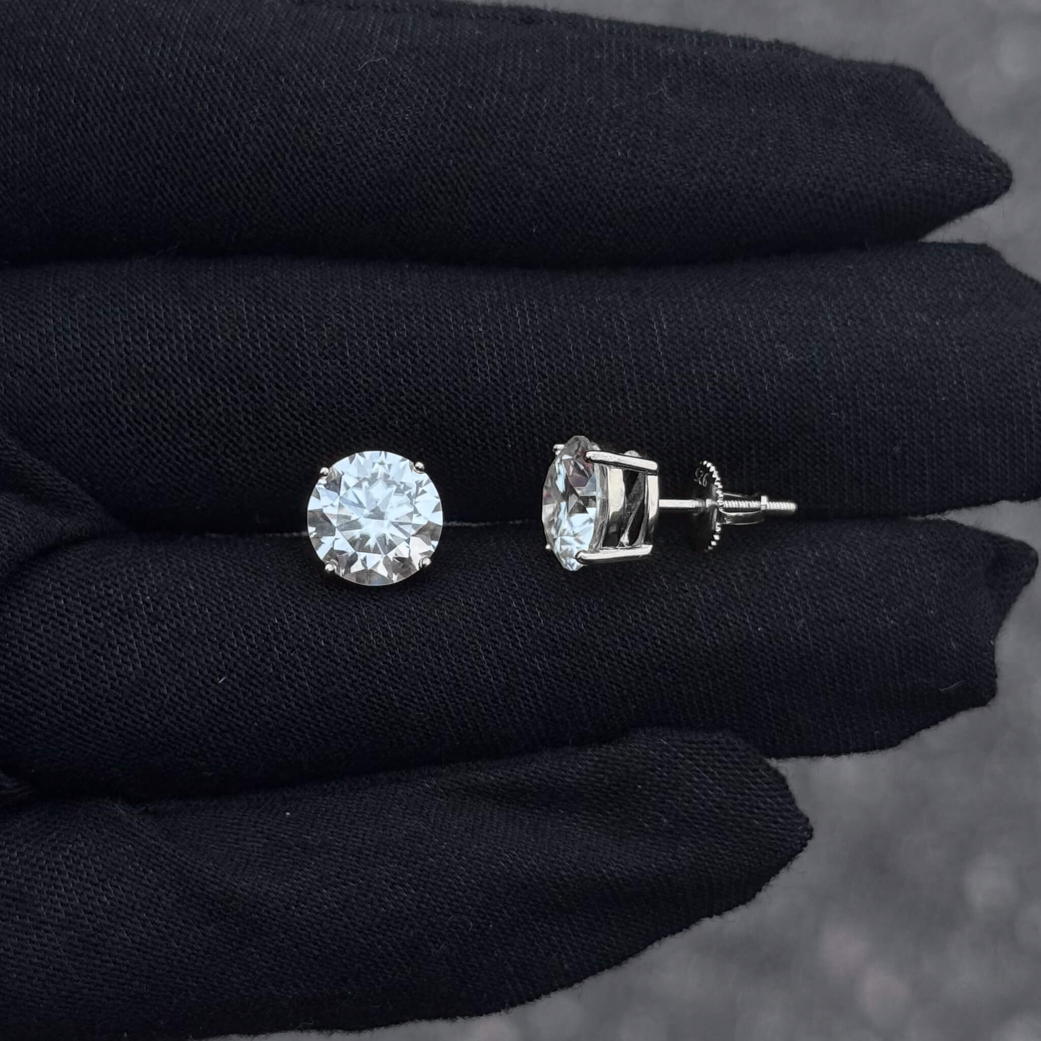 Round moissanite solitaire earrings with sterling silver screw-back posts shown on black glove.