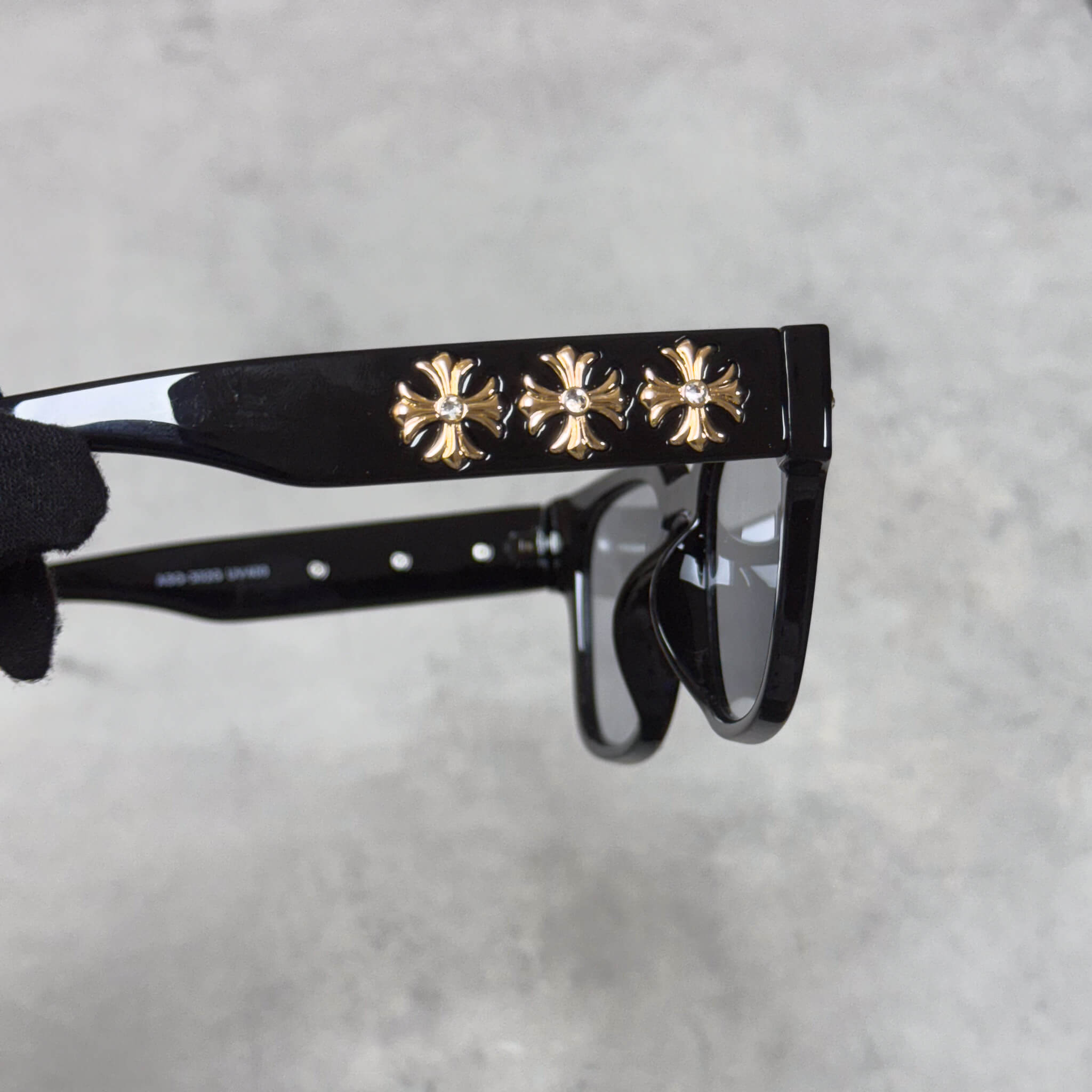 Close-up of black sunglasses arm showing three raised gold chrome cross emblems in a row.