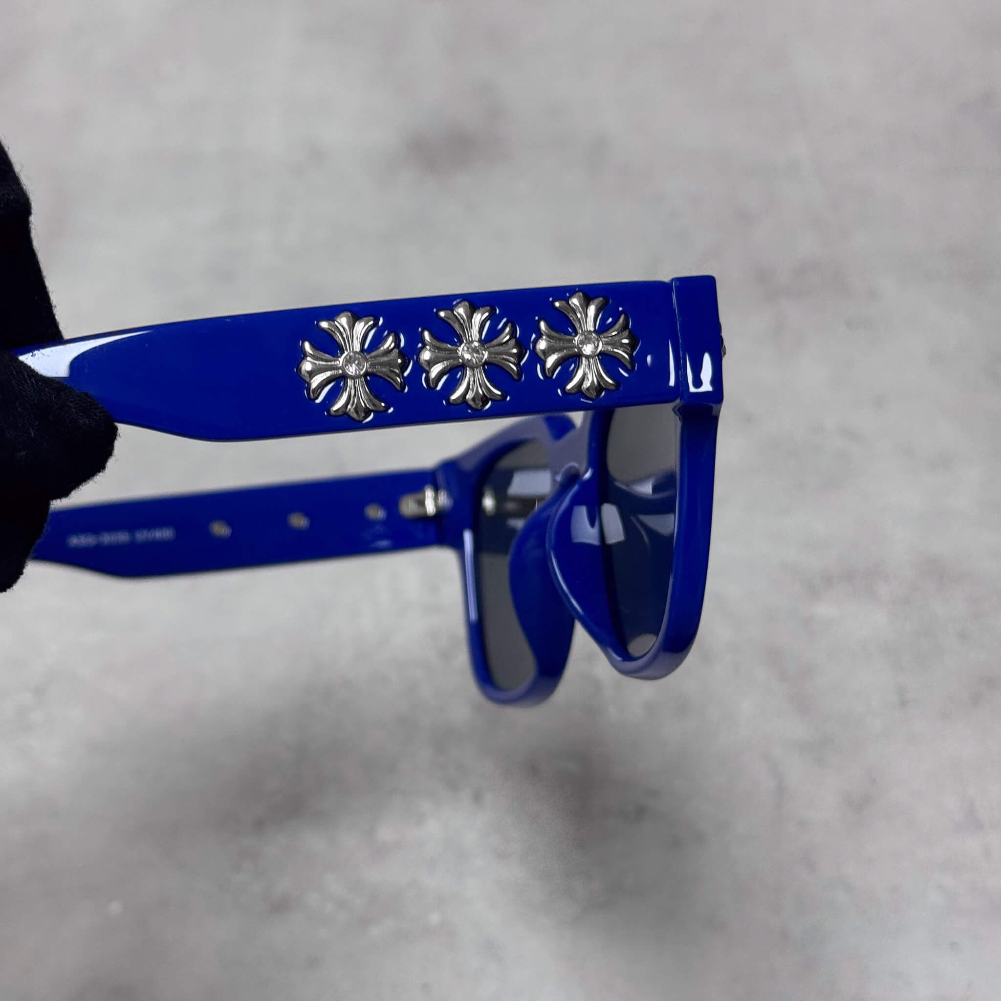Close-up of blue sunglasses arm showing three silver chrome cross emblems in polished detail.