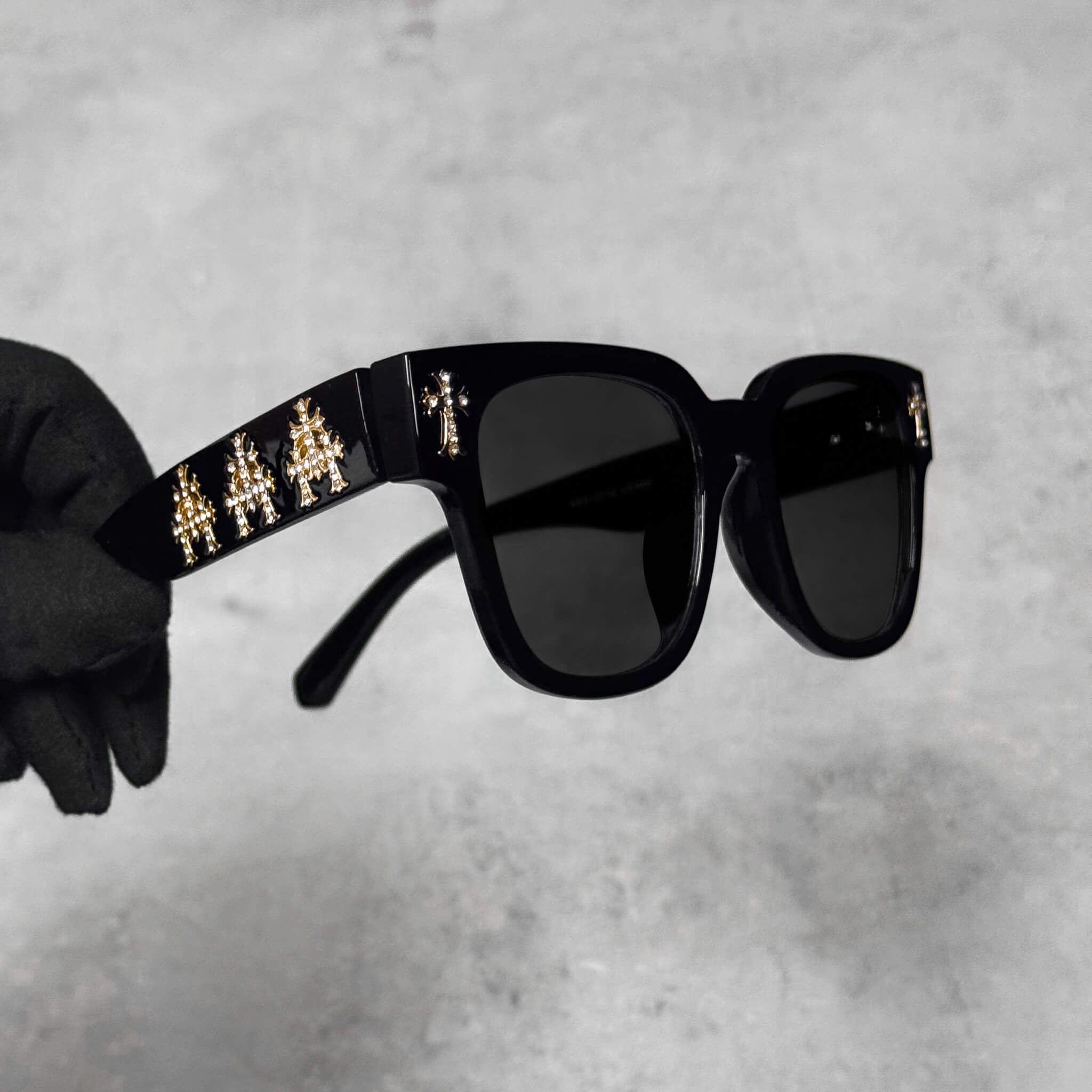 Angled view of black sunglasses featuring gold triple chrome crosses along the arms and temples.