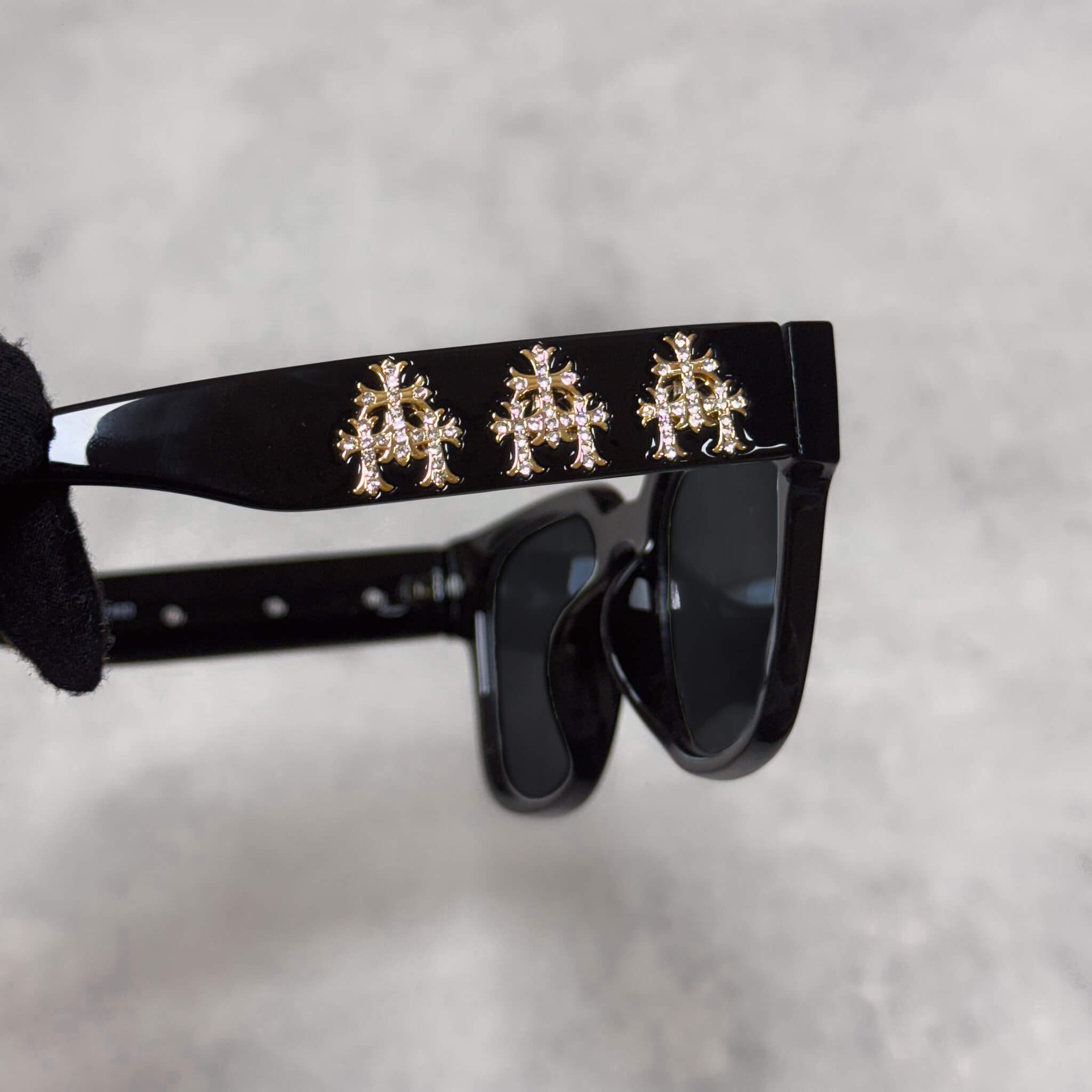 Close-up of black sunglasses arm showcasing three gold chrome cross emblems in polished detail.