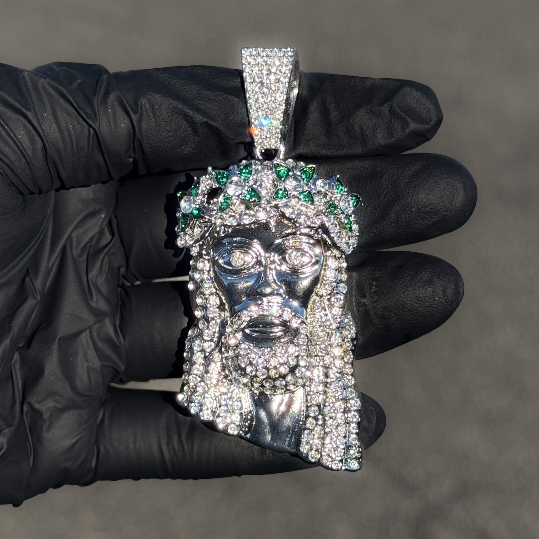 Silver iced-out Jesus pendant featuring green spiked crown and full pavé stones shown in hand.