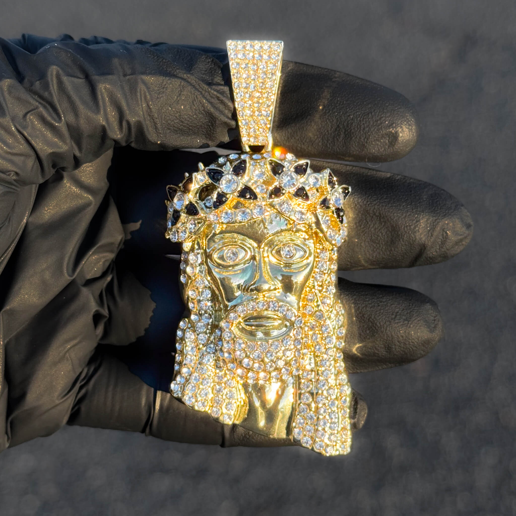 Gold iced-out Jesus spiked crown pendant with black stones displayed close-up in sunlight.