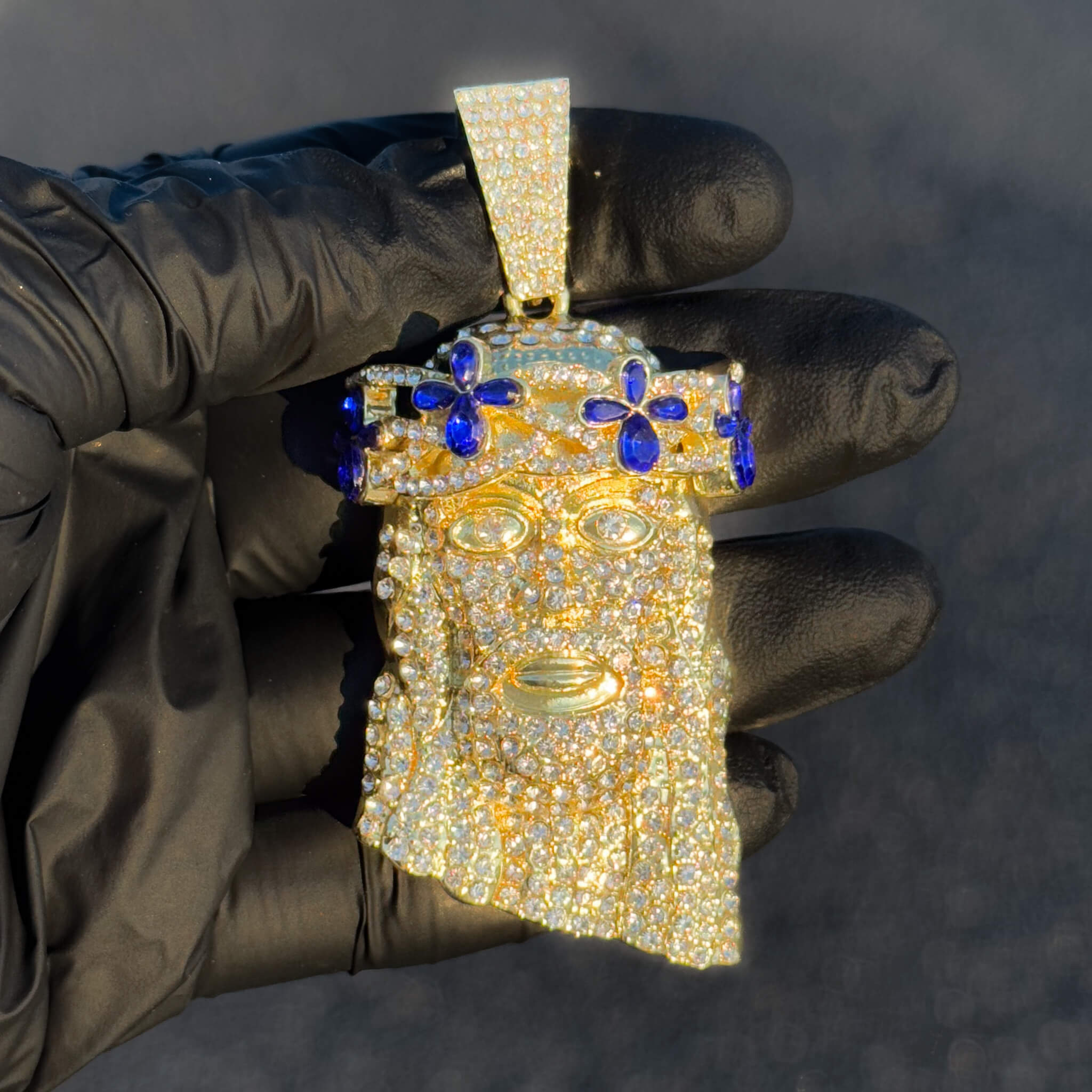 Large gold iced-out Jesus pendant with blue floral gemstones displayed in glove for close detail.