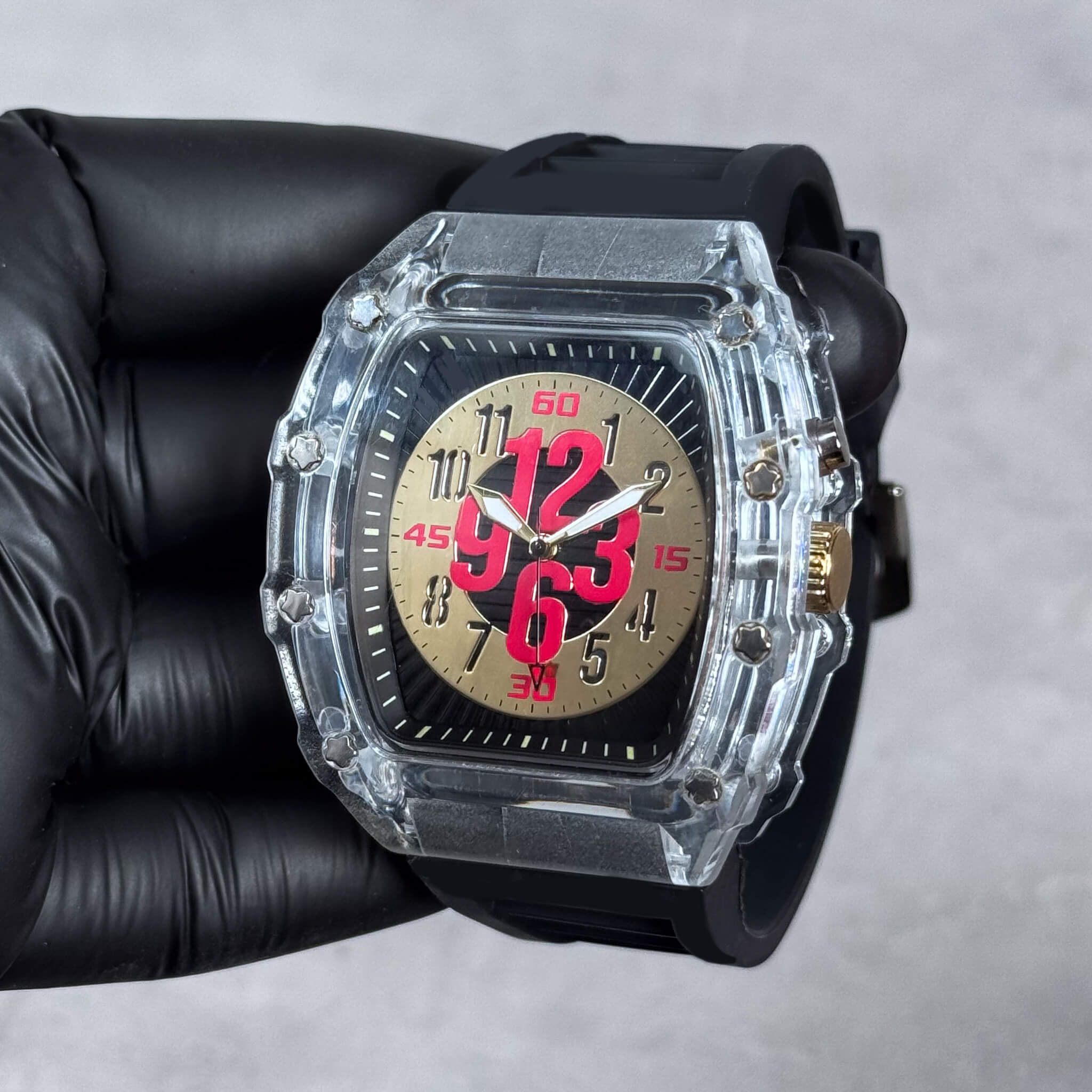 Gold-tone dial tonneau watch with oversized red numerals and clear case, black silicone strap.