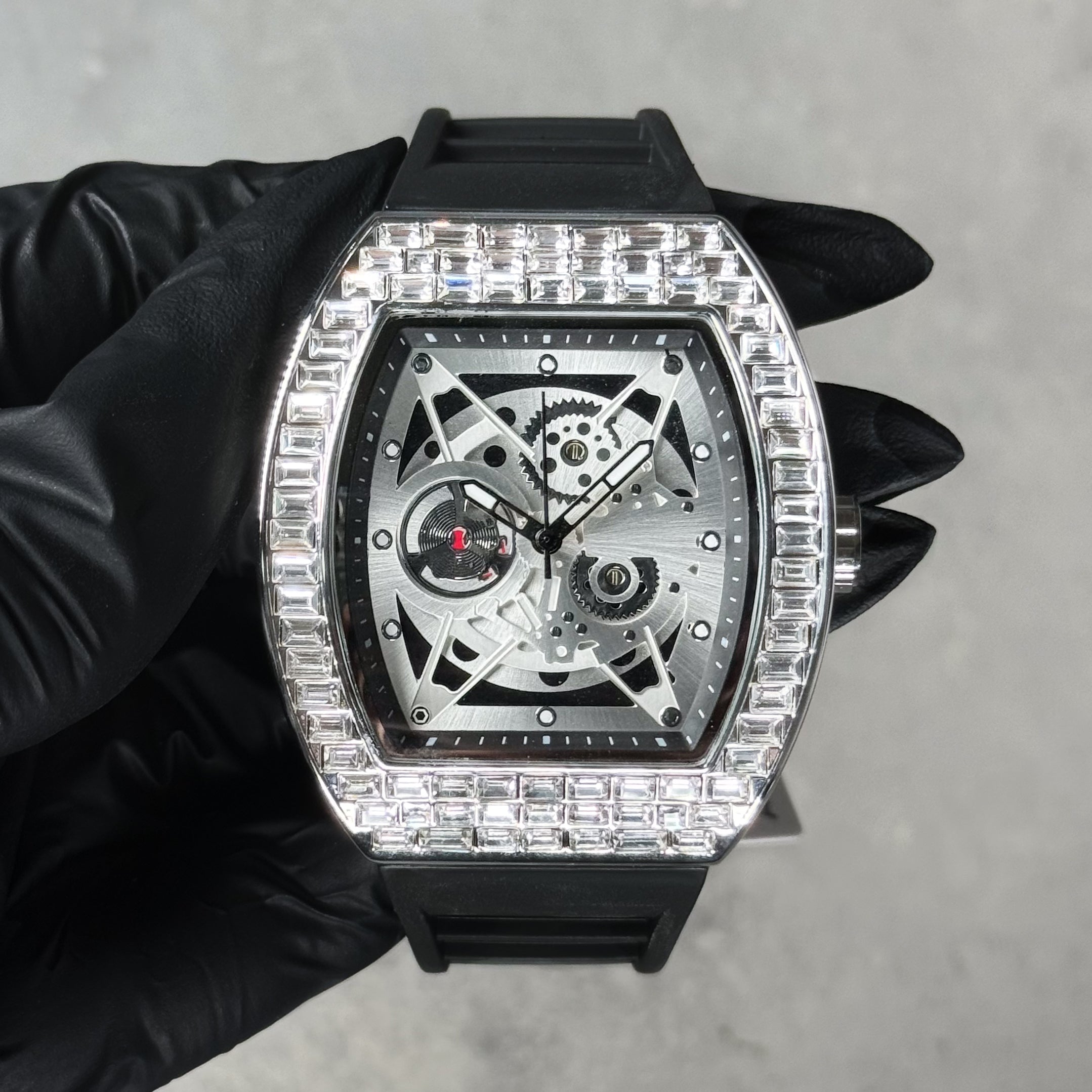 Silver men’s luxury iced watch with baguette crystal bezel, skeleton movement, and black silicone band.