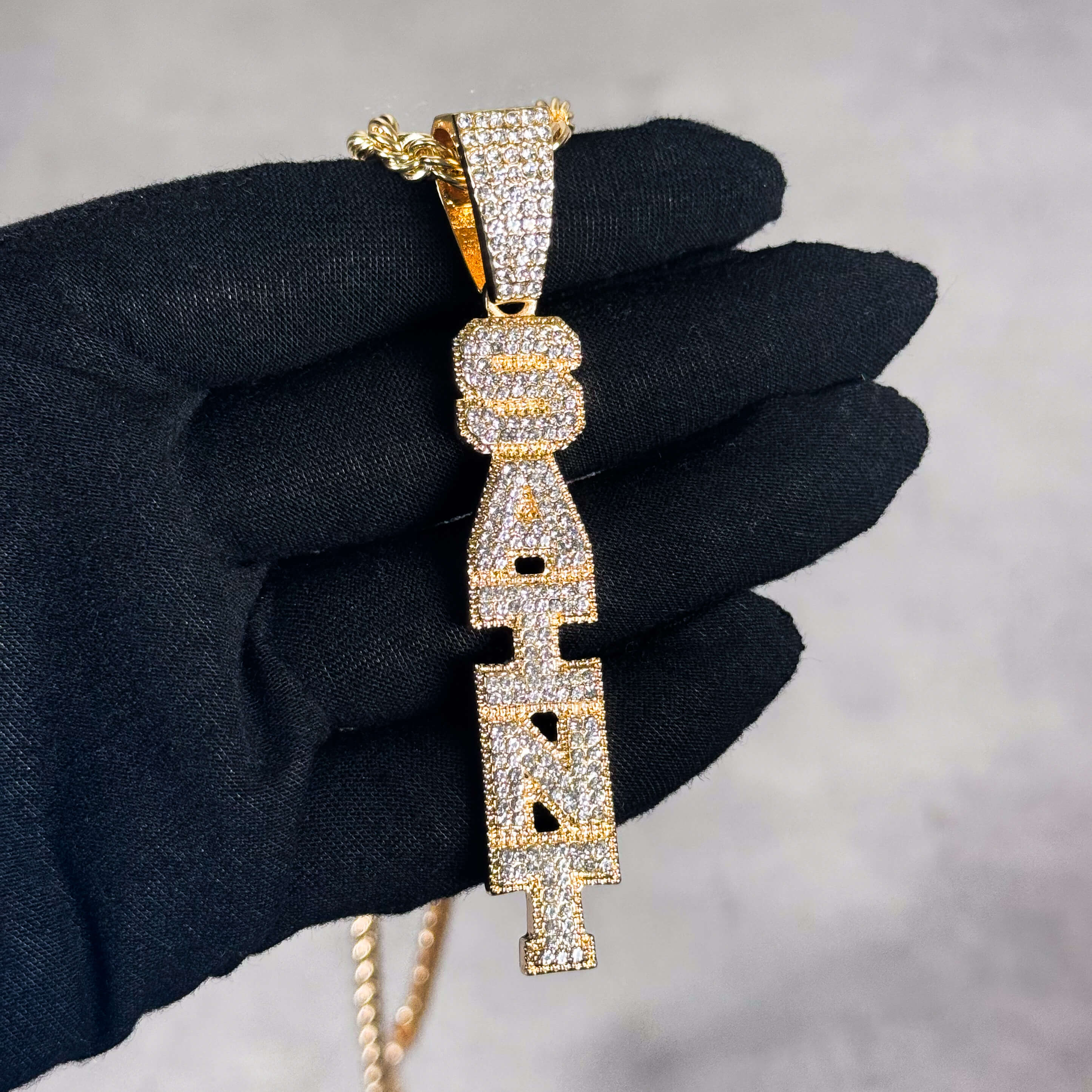 Gold Iced out Vertical Saint Pendant held in hand, pavé CZ stones and bold letter shape.
