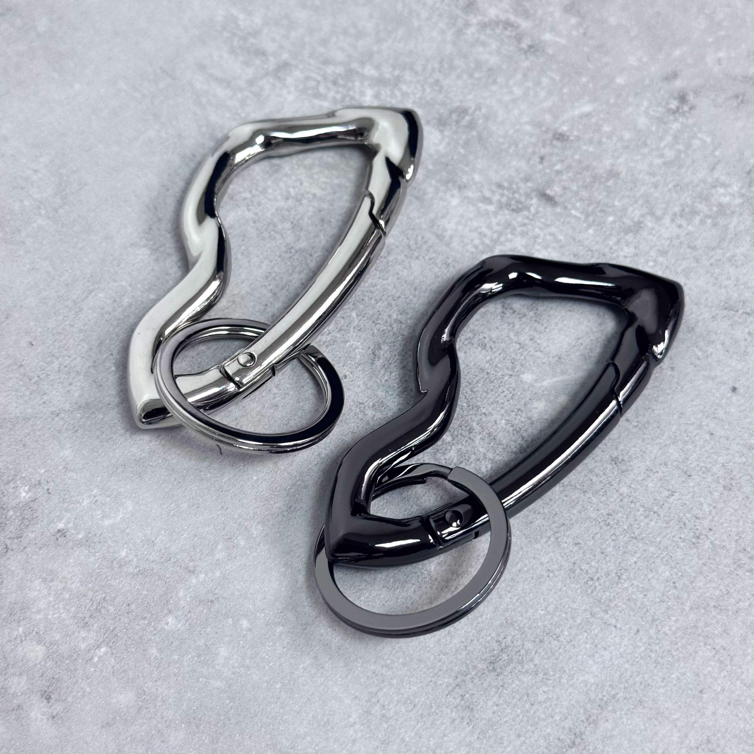 Irregular Y2K carabiner keychains in silver and black with wavy metal design on concrete surface.