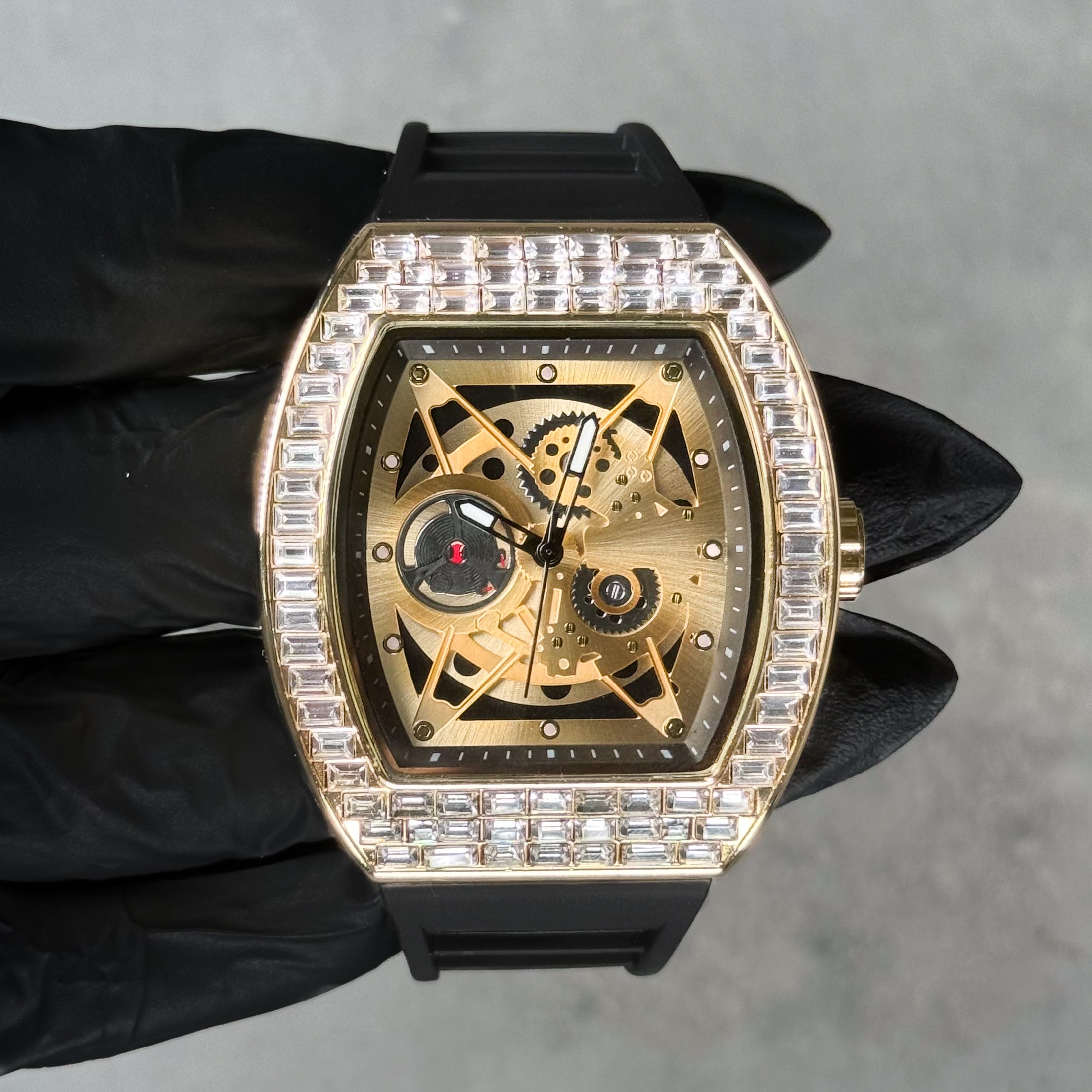 Gold men’s luxury iced watch with baguette crystals, skeleton dial, tonneau case, and silicone band.