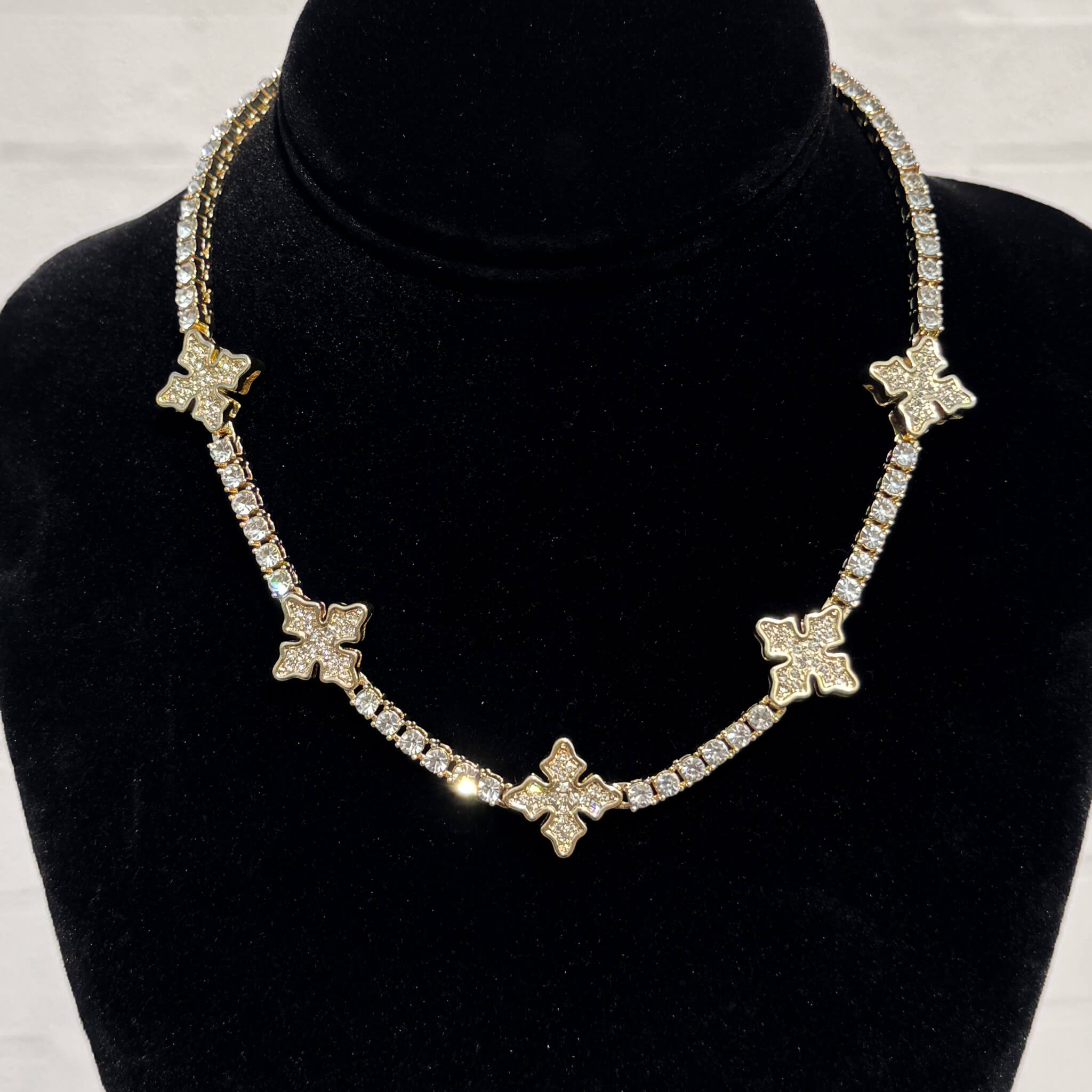 Iced Mini Cross Tennis Choker in gold with pavé crystal cross charms displayed on a black velvet stand.