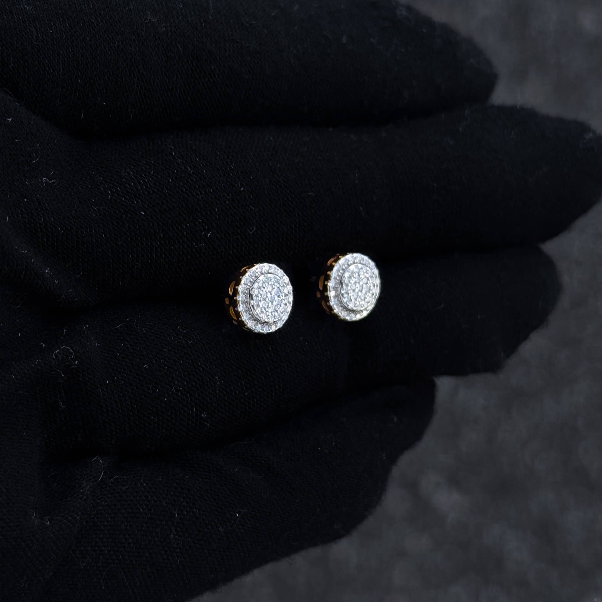 Small round moissanite halo stud earrings shown on black glove with gold-trim accents and pavé detail.