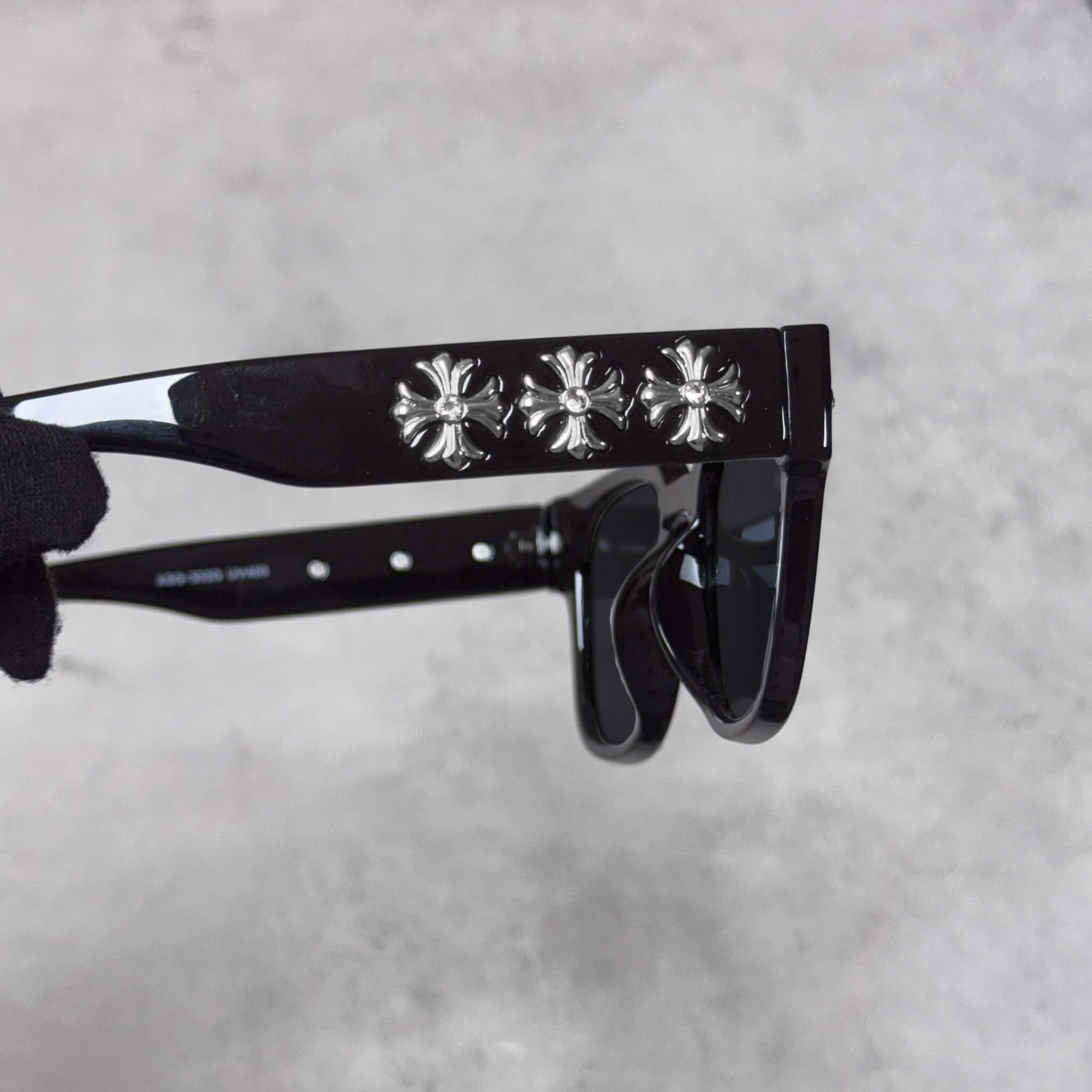 Close-up of black sunglasses with three silver chrome cross emblems along the temple arm.