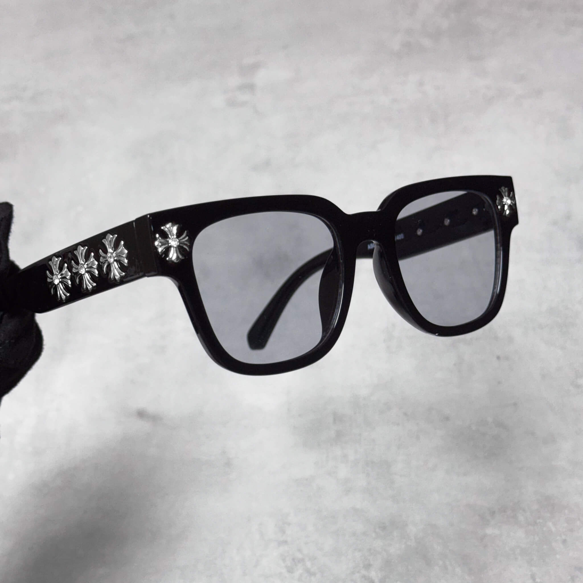 Side angle of black sunglasses featuring smoke lenses and silver chrome cross designs on temples.
