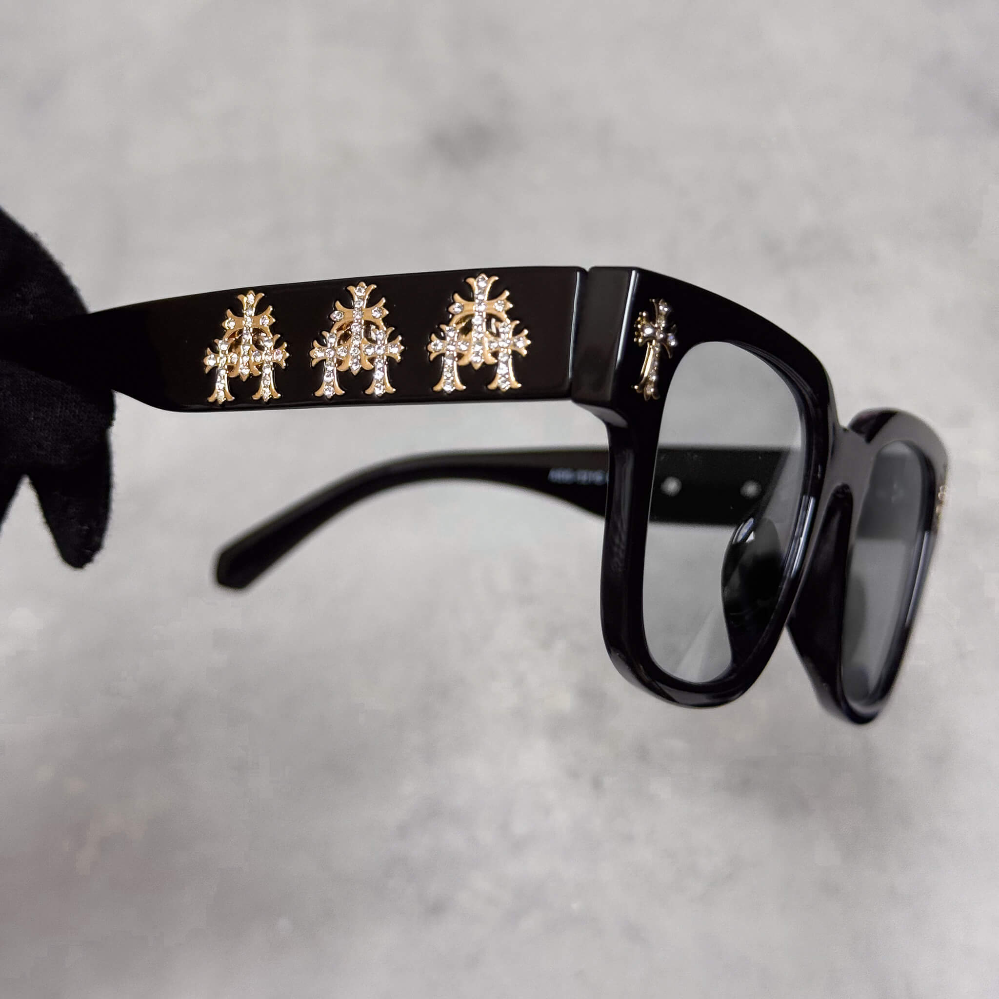 Close-up of black smoke-lens sunglasses showing gold triple-cross metal accents along temple arms.