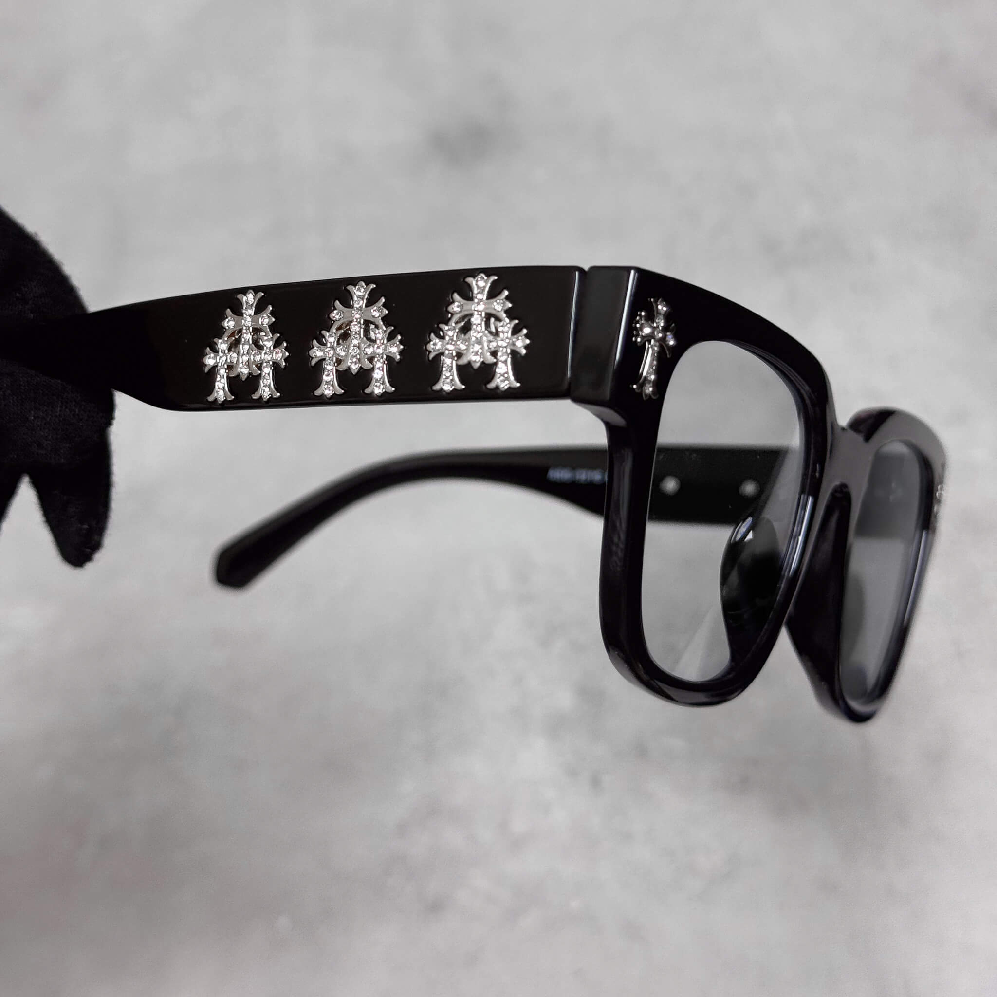 Close-up of black frame sunglasses highlighting silver triple-cross chrome embellishments on temples.