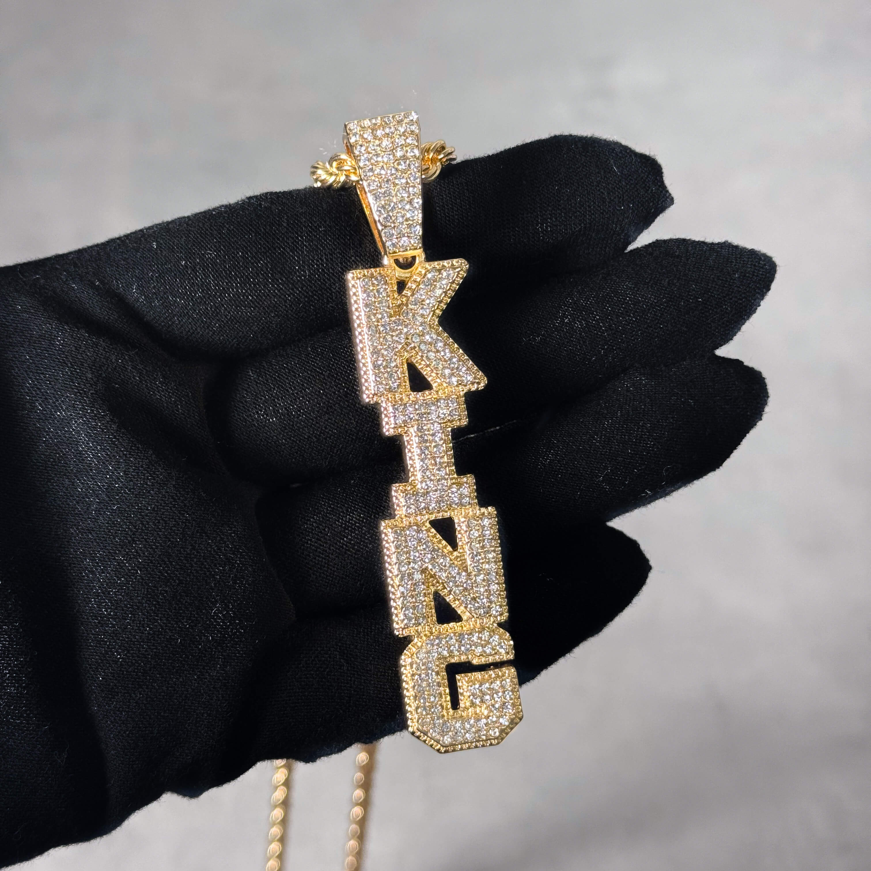 Gold Iced out Vertical King Pendant shown in hand, fully iced CZ letter design on rope chain.